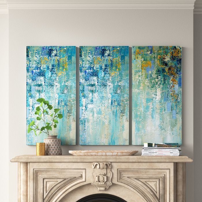 An abstract triptych painting hanging above a stone fireplace mantel, surrounded by a plant, candle, decorative tray, and stacked magazines