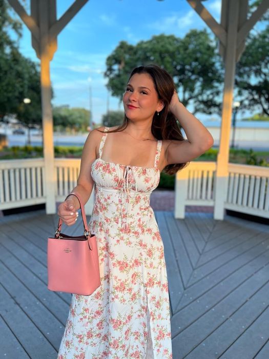 A reviewer posing in a floral dress with a pink handbag on a wooden gazebo