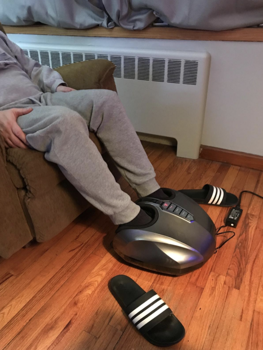 A reviewer enjoying the oval-shaped silver foot massager with two holes at the top where their feet are placed.