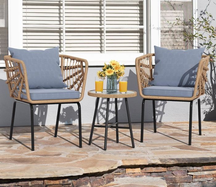 Two wicker chairs with light blue cushions and a small round table displaying yellow flowers and three glasses of orange juice on a patio, with no people in sight