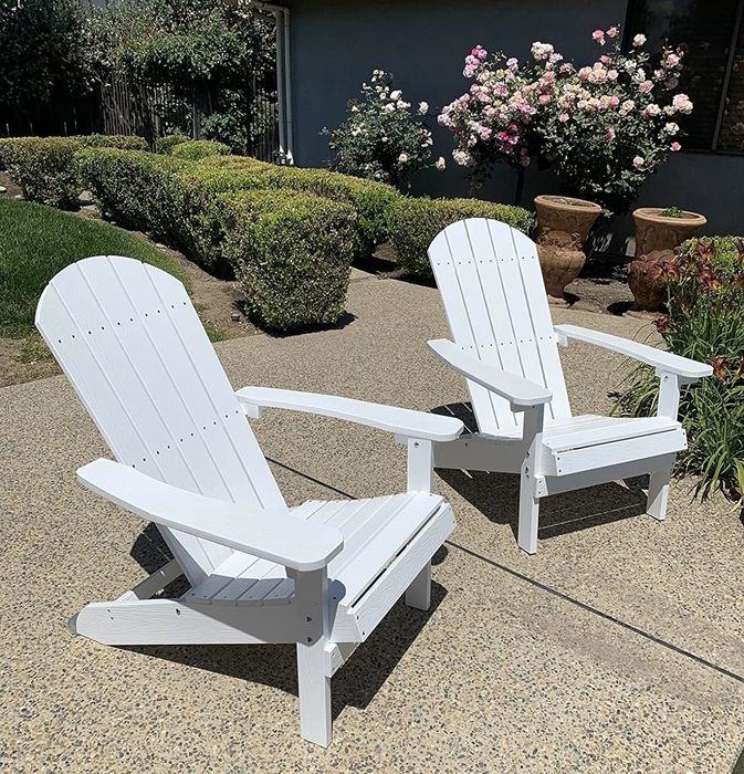 Two white Adirondack chairs are placed on a concrete patio in front of a house, surrounded by hedges and blooming flowers