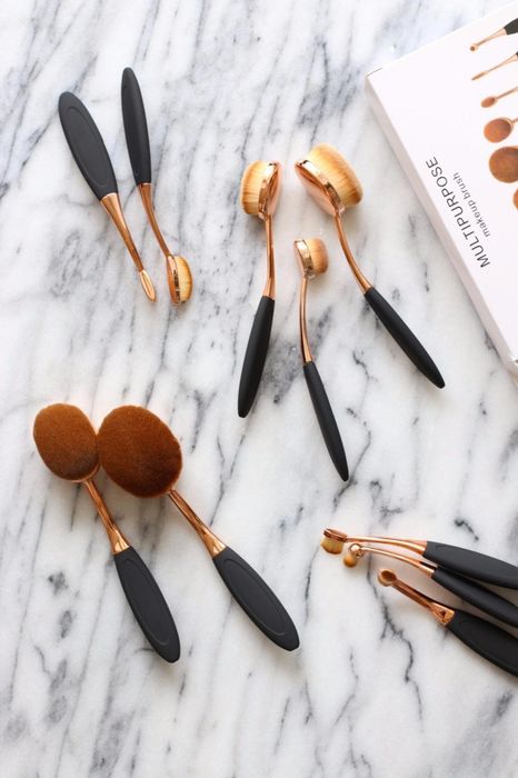 A set of oval makeup brushes with sleek black handles and gold accents, elegantly displayed on a marble surface next to a white box labeled.