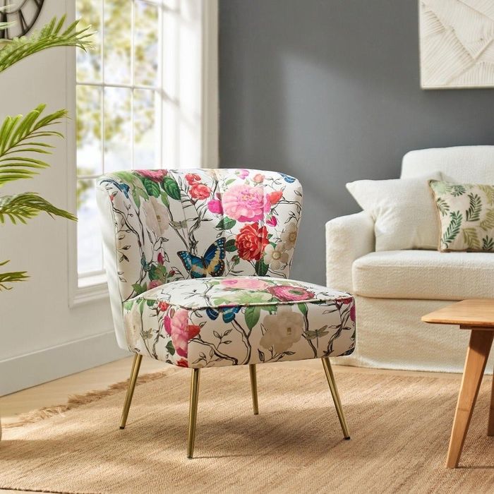 A floral accent chair with gold legs, placed in a living room alongside a white sofa, a wooden coffee table, and a potted plant.