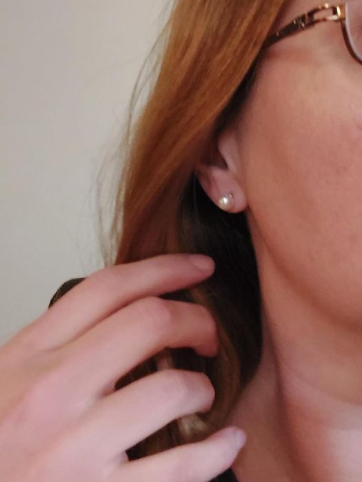 A close-up shot of a person’s ear adorned with a small pearl earring, with a hand (light-skinned) near the ear, and the person wearing glasses