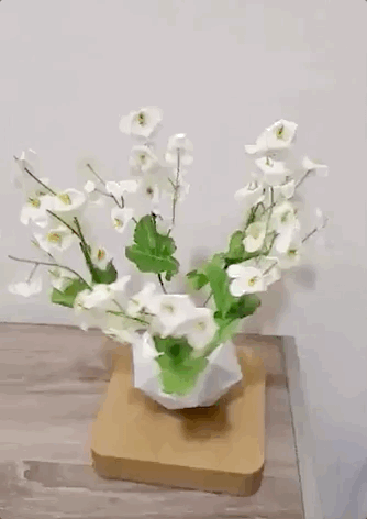 A gif of a planter with white blossoms floating above a light-colored wooden base