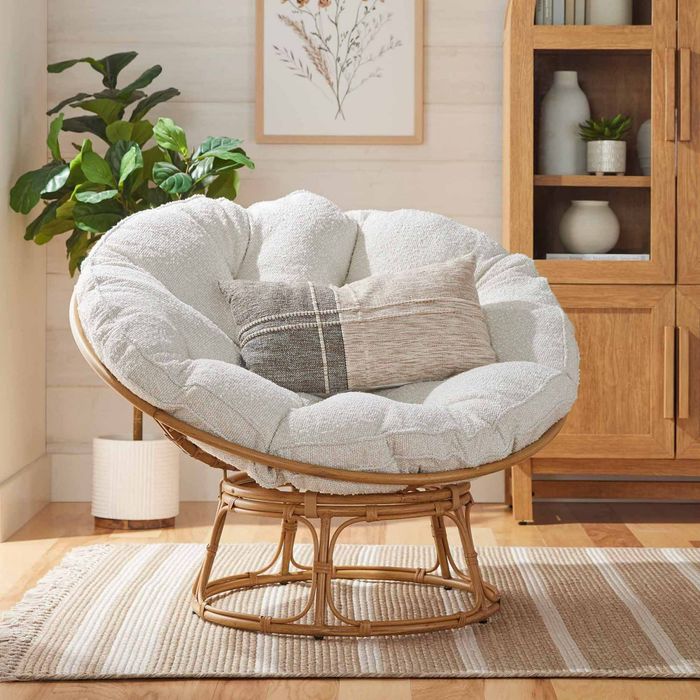 A round, plush chair with a thick cushion and a striped pillow, sitting atop a patterned rug, surrounded by plants and wooden furniture in a cozy living room.