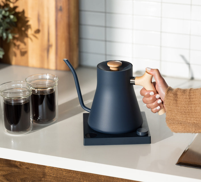 A hand reaching for the blue and maple wood electric kettle resting on a countertop