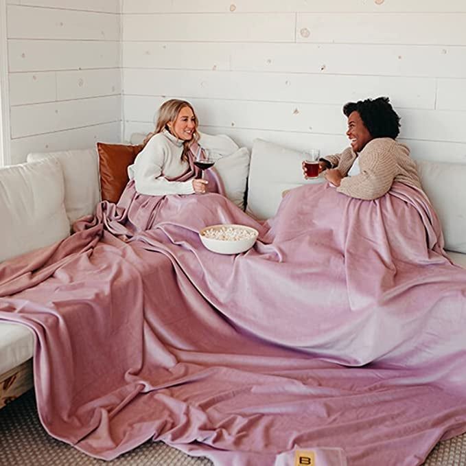 Two models comfortably snuggled together beneath a large pink blanket.