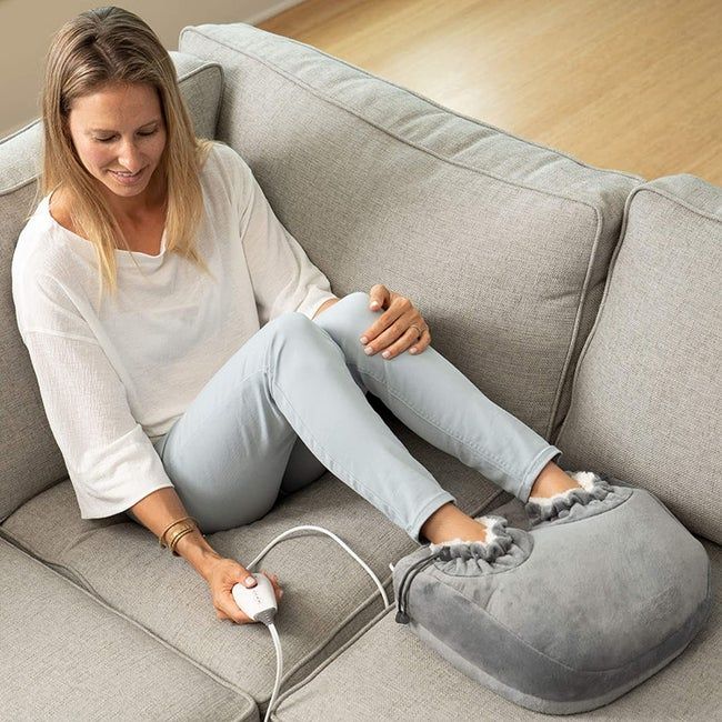 A person relaxing on a sofa with their feet nestled inside a large, plush foot heater, each foot secured by a hole and cinched to ensure extra coziness.
