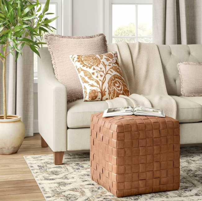 A woven leather ottoman in a cozy living room, paired with a beige sofa adorned with a floral cushion