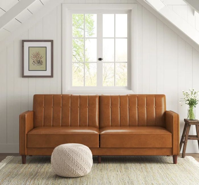 rectangular brown faux leather couch in a spacious, open area next to a small pouf