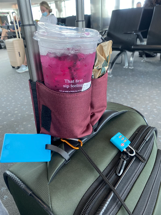 A drink caddy clipped to a suitcase handle, holding a Starbucks cup and a sandwich