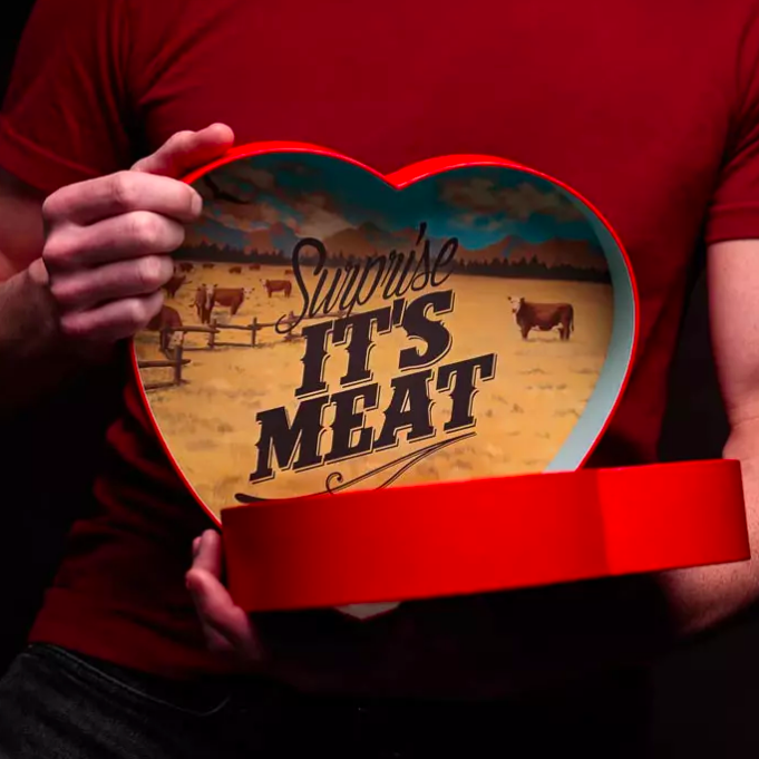 A heart-shaped box with the lid open, revealing jerky inside and a playful message on the bottom