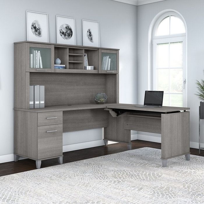 L-shaped gray desk with built-in storage shelves and drawers, topped with a laptop, set in a bright room with large windows and decorative wall art