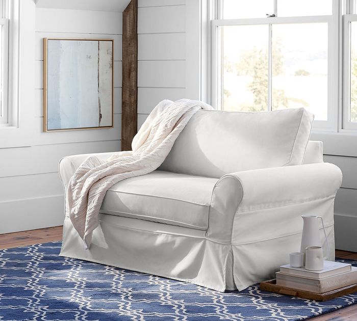 A white slipcovered chair draped with a beige throw blanket, set in a room with a blue patterned rug