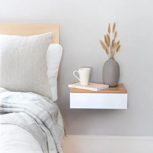 A rectangular white wood drawer with a light brown wood top, attached to the wall next to the bed. It's holding a book, coffee cup, and a vase.