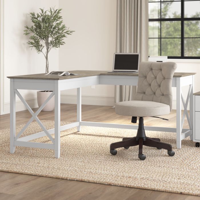 L-shaped desk with a laptop and notebook, placed on a carpet. A comfy cushioned rolling chair sits nearby in a minimalist home office setup