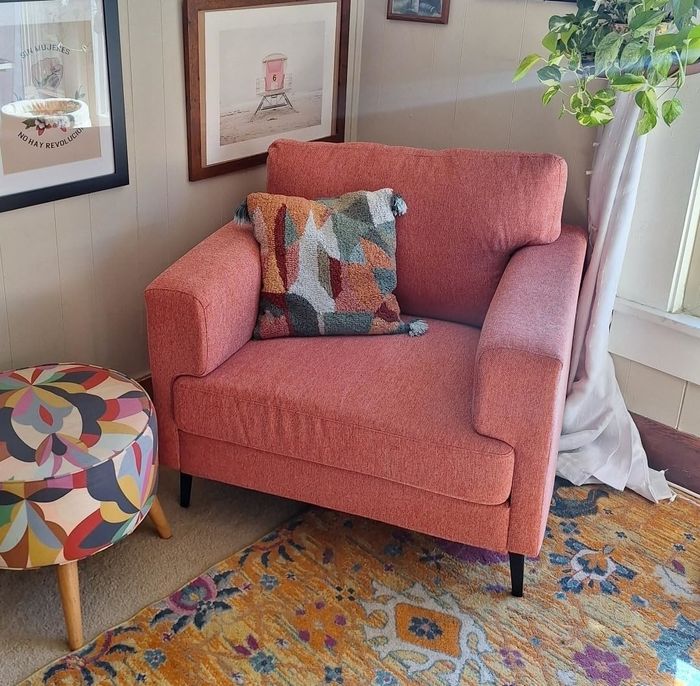 A cozy corner in a living room featuring a peach armchair, a colorful pillow, a round patterned ottoman, and framed artwork on the walls, beside a window adorned with a plant.