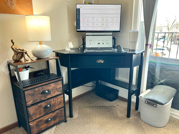 A home office setup with a small black desk, laptop, monitor, lamp, and a side table with drawers. A balcony view is visible, with various personal items neatly arranged