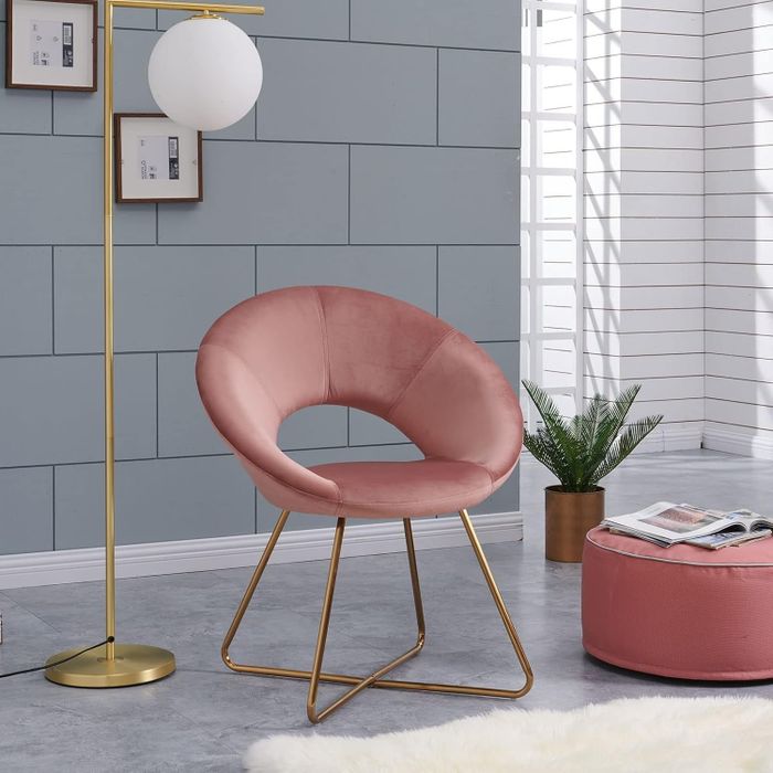 A chic living area featuring a pink velvet chair with gold legs, a matching ottoman topped with magazines, a gold floor lamp, framed pictures, and a decorative plant.