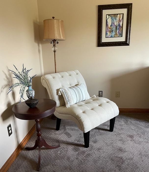 An elegant tufted armchair in beige upholstery, positioned next to a wood side table with decorative items and a floor lamp