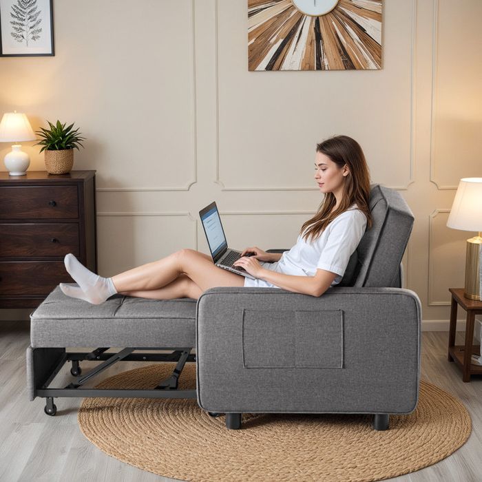 A woman relaxing on a gray convertible sofa bed, working on a laptop in a cozy, well-lit living room with modern decor
