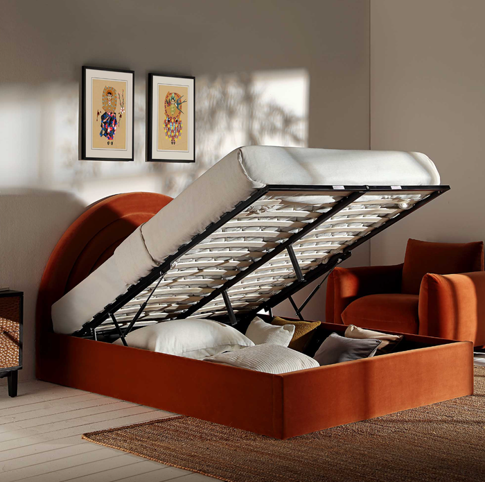 A storage bed with the mattress raised to reveal a spacious compartment, set in a modern room decorated with geometric prints and a plush chair in the background.
