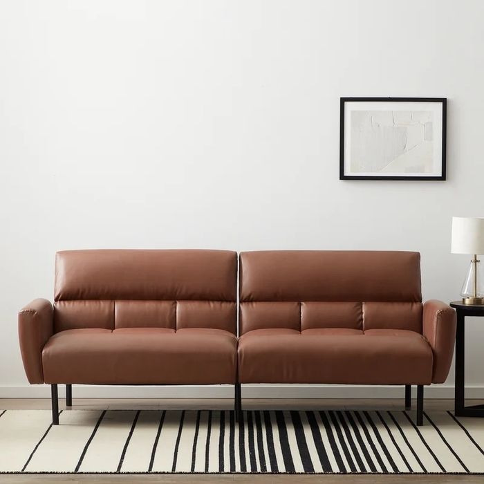 A modern brown sofa with a minimalist design is placed in a living room featuring framed abstract art, a lamp, and a striped rug