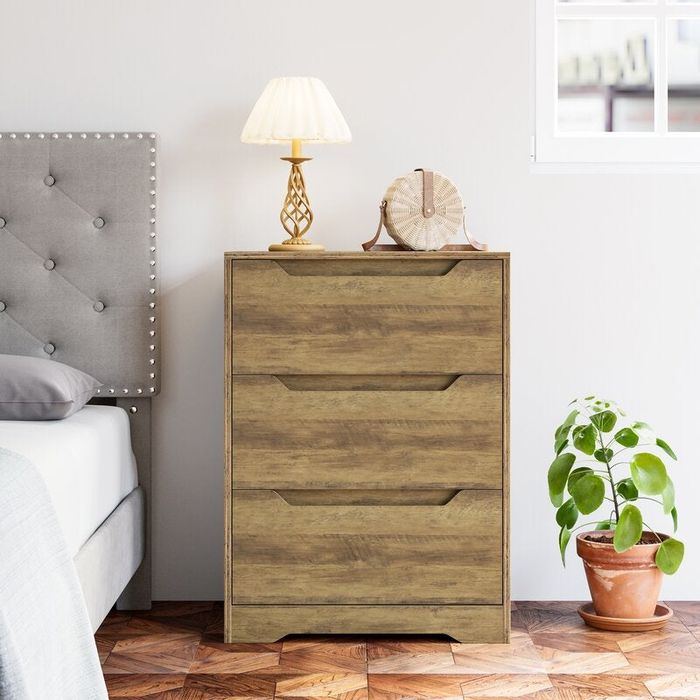 A brown three-drawer dresser placed beside a bed