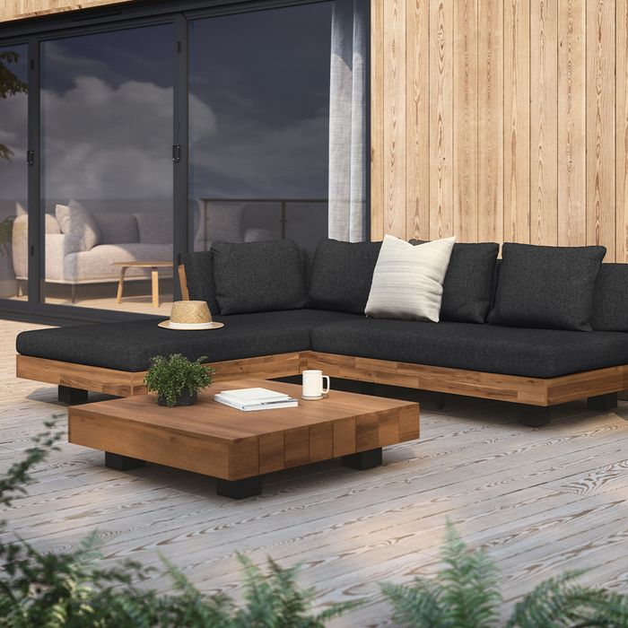 A wooden outdoor patio set featuring a dark cushioned L-shaped sofa, a low square coffee table, and potted plants, arranged on a deck with glass doors visible in the background.
