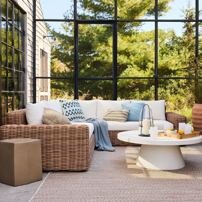 A cozy outdoor patio featuring a wicker sofa, plush pillows, a round coffee table, and decorative accents, all set in a bright garden room.