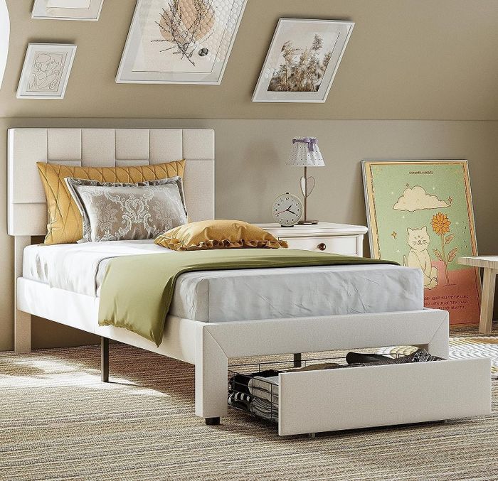 A chic bedroom featuring a single bed with under-bed storage drawers, accompanied by decorative wall art, a bedside table, and a cozy arrangement of plush pillows.