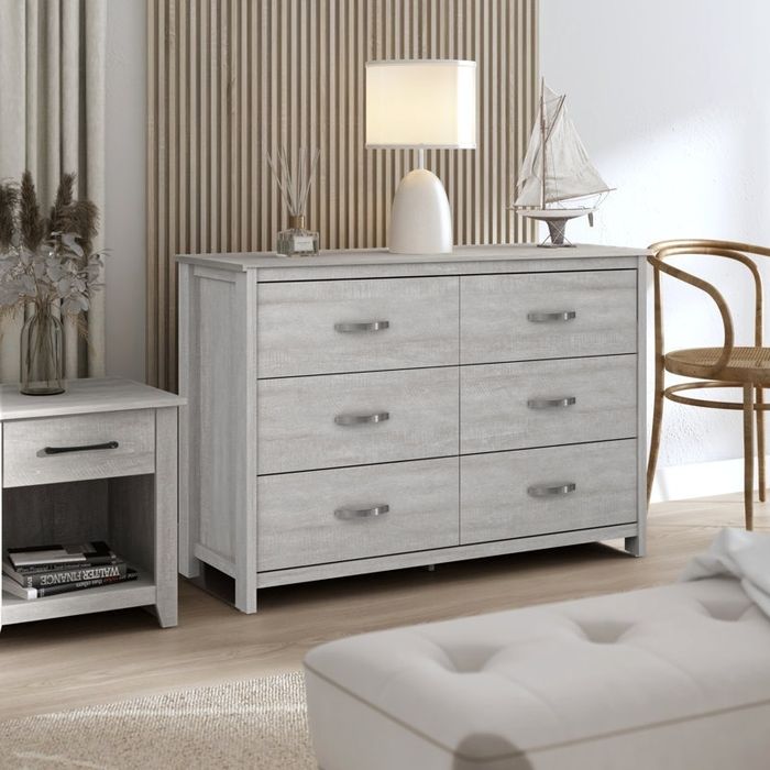 A modern bedroom setup featuring a light wood dresser, a matching nightstand, and a lamp. The decor includes dried flowers and a model sailboat.