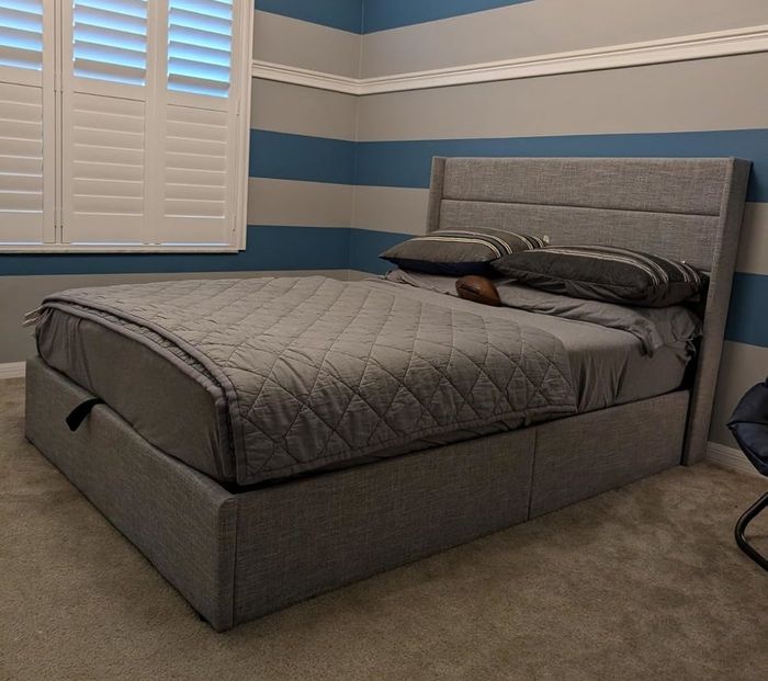 A sleek modern bed with grey bedding neatly arranged, positioned against a wall with broad horizontal stripes. The window to the left is covered with white shutters.
