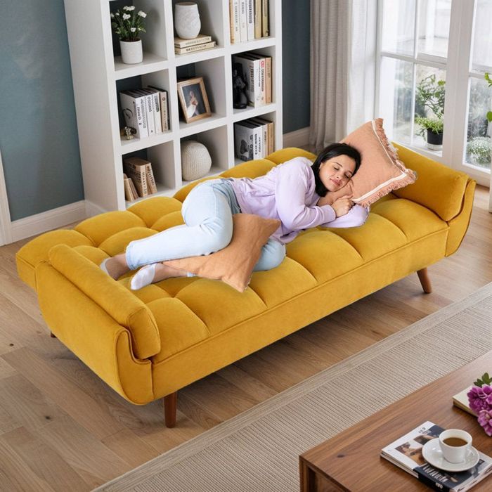 A person relaxing on a mustard-yellow chaise lounge, holding a peach-colored pillow, in a cozy living room filled with bookshelves and natural light.