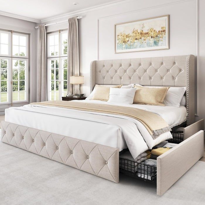 A luxurious beige upholstered bed placed in a bright, airy bedroom, featuring practical under-bed storage drawers. The room is decorated with picture frames and large windows in the background.