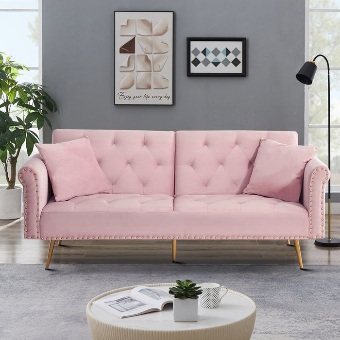 A modern pink tufted sofa with pillows, placed in a cozy living room setting, complemented by wall art and a floor lamp for added decor.