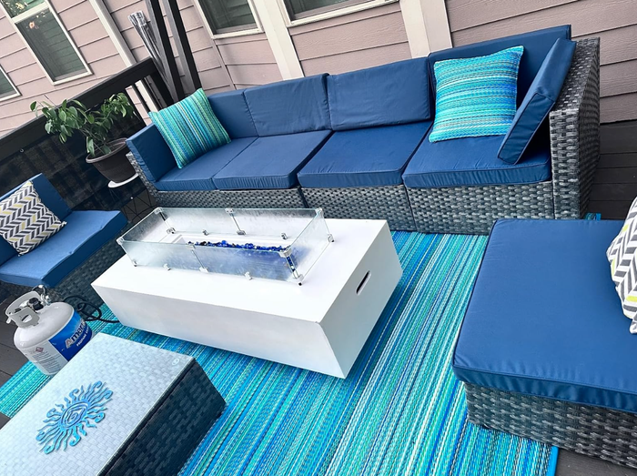 An outdoor seating area showcasing rattan furniture with blue cushions, decorative patterned pillows, a white fire pit, and a propane tank.