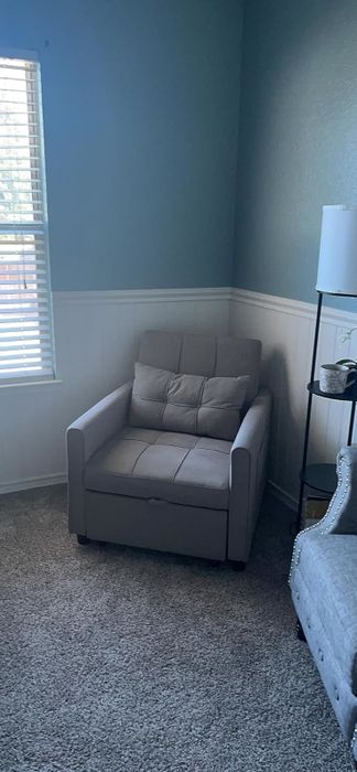 A cozy upholstered armchair nestled in a corner of a room, with a window, lamp, and shelves in view, creating the perfect reading or relaxation spot.