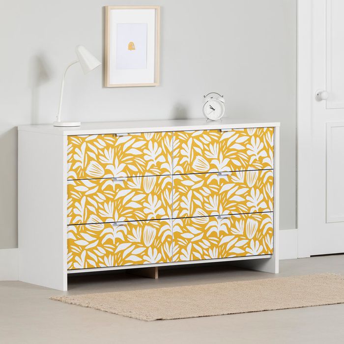 A white dresser with three drawers, each featuring a yellow and white floral pattern. A white lamp and clock rest on top, with a framed picture hung on the wall above.