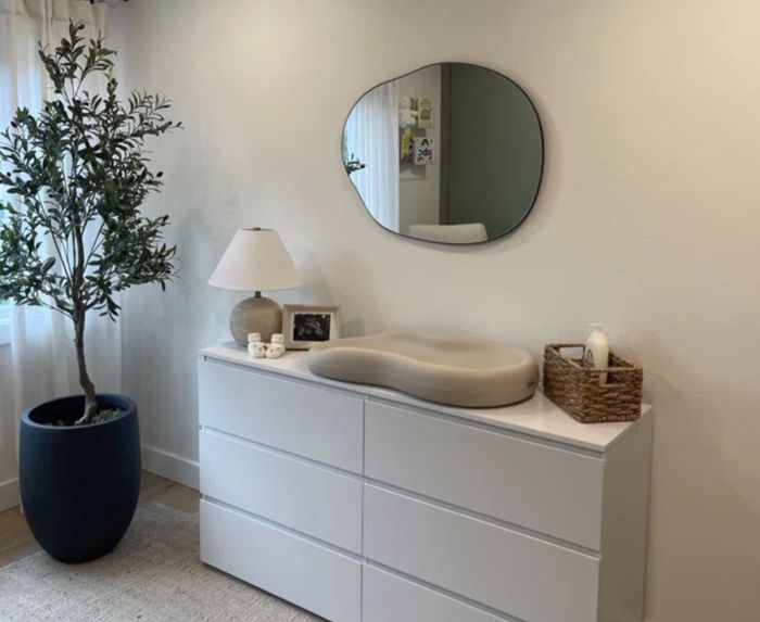 Modern bedroom dresser featuring a wavy mirror, potted plant, basket, and lamp