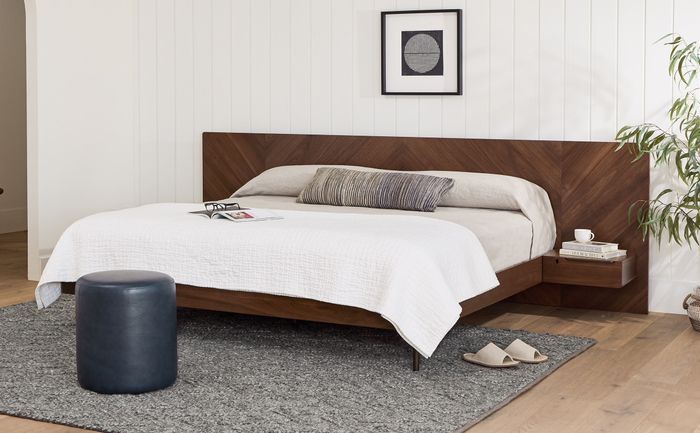 A minimalist bedroom with a sleek bed, a bedside table, and a round ottoman, with a magazine placed on the bed.