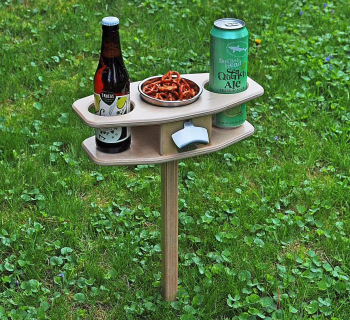 A compact table mounted on a stake with two cupholders, a center compartment filled with snacks, and an attached bottle opener.