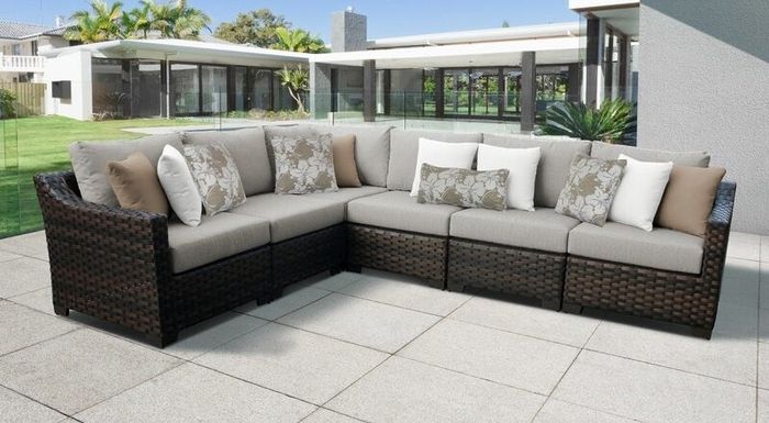 An outdoor wicker sectional sofa set with beige cushions and decorative patterned pillows, placed on a spacious patio with modern architecture and lush greenery in the background.