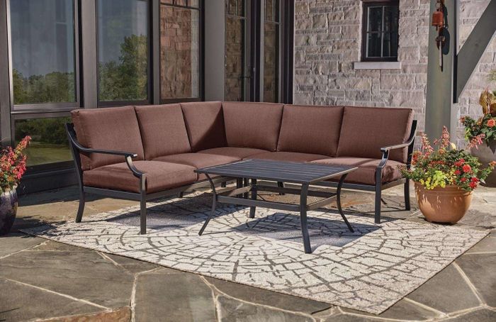An outdoor patio showcasing a brown cushioned metal sectional sofa paired with a matching rectangular coffee table, placed on a patterned rug and surrounded by lush potted plants.