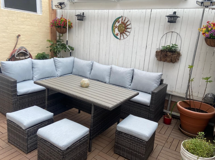 A patio set featuring a wicker bench with light cushions, a rectangular table, and matching stools, arranged in a charming garden space adorned with hanging plants and wall decor.