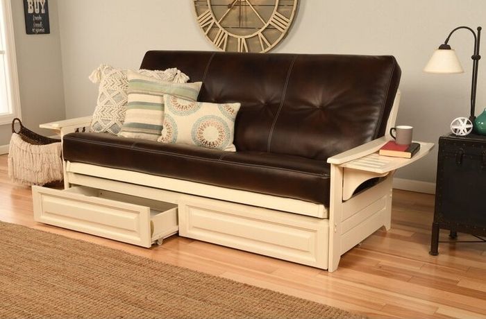 A chic futon with sleek black leather cushions, complemented by striped and patterned pillows, and equipped with wooden storage drawers underneath. A lamp and clock set the scene in the background.