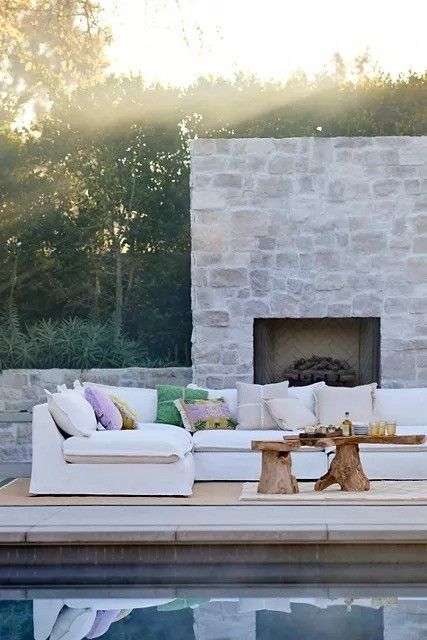 An outdoor patio featuring a white sectional sofa adorned with colorful pillows and rustic wooden tables, situated by a pool with a stone fireplace in the background.