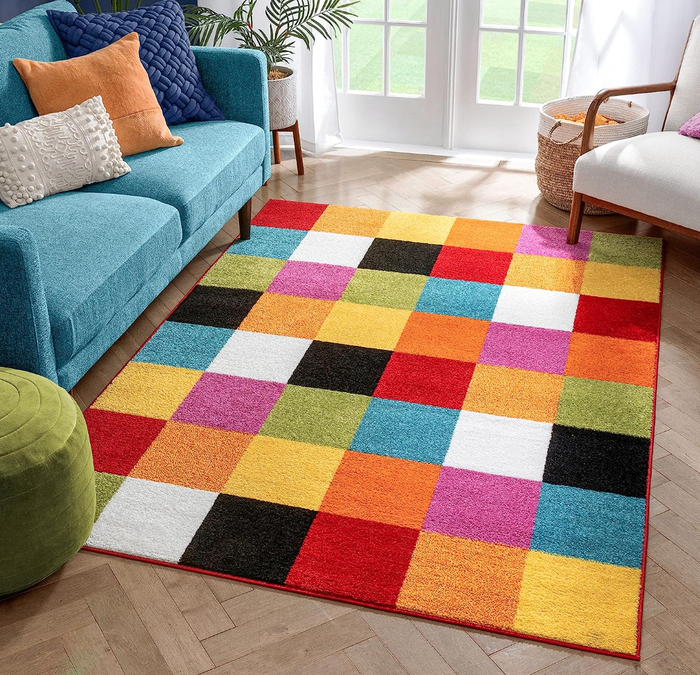 A colorful checkered rug with varying shades, placed in a cozy living room with a blue couch, a green ottoman, and a large window.