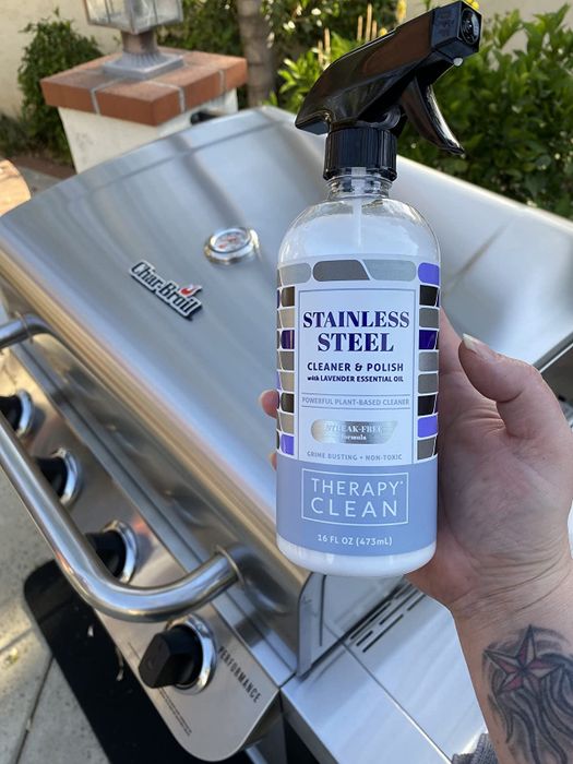 A reviewer holding the bottle of stainless steel cleaning spray with their stainless steel grill in the background